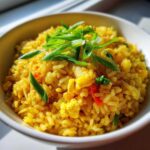 A close-up of a white bowl filled with bright yellow Gluten-Free Lemon Pepper Fried Rice, topped with fresh green scallions.