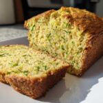 A golden-brown loaf of Gluten-Free Lemon Zucchini Bread, with two slices cut and resting against the main loaf on a white plate.