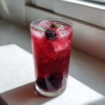 A tall glass filled with vibrant red Iced Blackberry Sage Tea, ice cubes, fresh blackberries, and sage leaves.