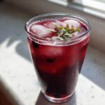 A tall glass of deep red Iced Blackberry Sage Tea filled with ice and garnished with fresh sage leaves.