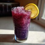 A tall glass filled with a vibrant, icy Iced Blueberry Lemonade Slush, garnished with a bright lemon slice.