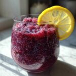A close-up of a vibrant, deep purple Iced Blueberry Lemonade Slush garnished with a bright lemon slice.