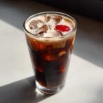 A tall glass filled with Iced Cherry Vanilla Cold Brew, ice, creamy foam, and a bright red cherry garnish.