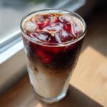 Close-up of a tall glass filled with Iced Cherry Vanilla Cold Brew, showing layers of milk, dark coffee, and a red cherry syrup swirl over ice.