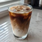 Close-up of a tall glass containing an Iced Honey Vanilla Latte with Oat Milk, showing layers of dark espresso and white milk over ice.