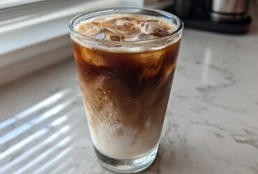 Close-up of a tall glass containing an Iced Honey Vanilla Latte with Oat Milk, showing layers of dark espresso and white milk over ice.