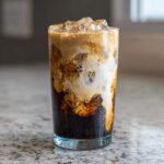 A tall glass filled with an Iced Honey Vanilla Latte with Oat Milk, showing dark coffee swirling with creamy white oat milk and ice cubes.