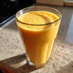 A tall glass filled with a thick, bright orange Iced Orange Creamsicle Smoothie sitting on a granite countertop.