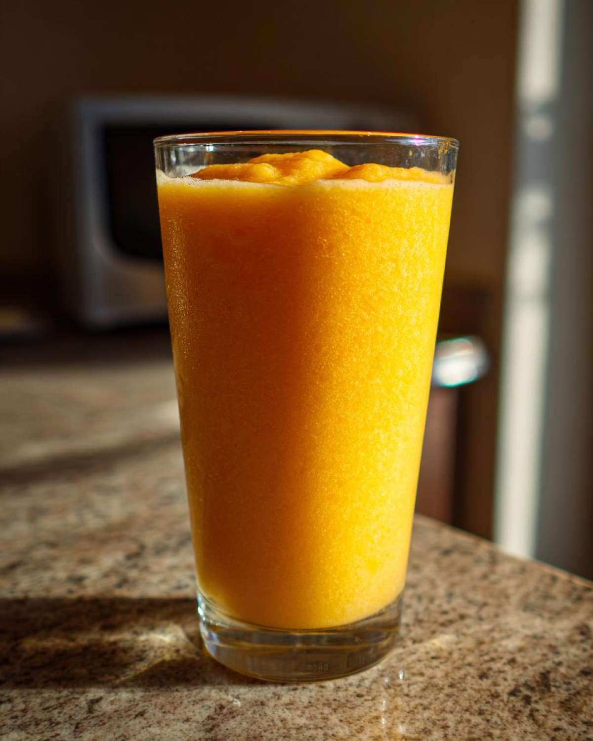 A tall glass filled with a thick, bright orange Iced Orange Creamsicle Smoothie sitting on a speckled countertop.
