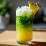 A tall glass filled with an Iced Pineapple Matcha Spritz, showing yellow pineapple juice layered beneath bright green matcha, topped with ice and a pineapple wedge.