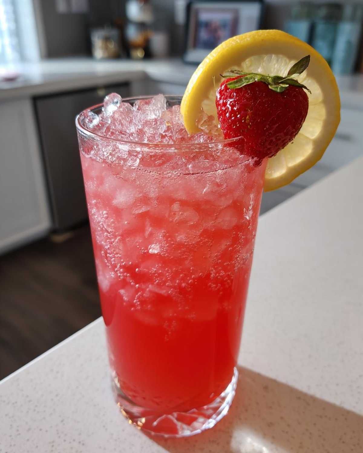 A tall glass filled with bright red Iced Strawberry Hibiscus Tea Lemonade, topped with crushed ice and garnished with a lemon slice and fresh strawberry.