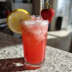 A tall glass of bright red Iced Strawberry Hibiscus Tea Lemonade garnished with a lemon slice and fresh strawberry.