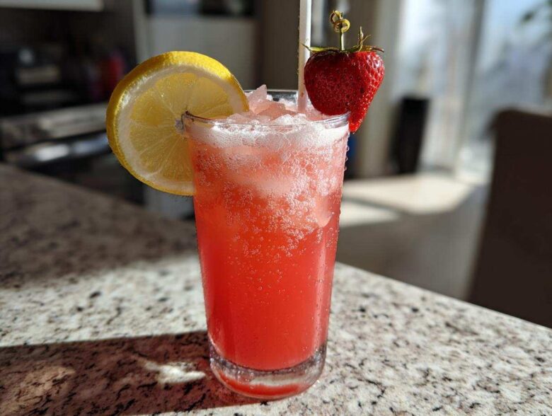 A tall glass of bright red Iced Strawberry Hibiscus Tea Lemonade garnished with a lemon slice and fresh strawberry.