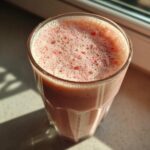 Close-up of a frothy Iced Vanilla Strawberry Protein Shake in a glass, catching the sunlight.