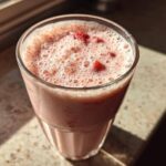 Close-up of a frothy Iced Vanilla Strawberry Protein Shake topped with small strawberry pieces in a clear glass.
