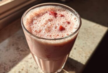 Close-up of a frothy Iced Vanilla Strawberry Protein Shake topped with small strawberry pieces in a clear glass.