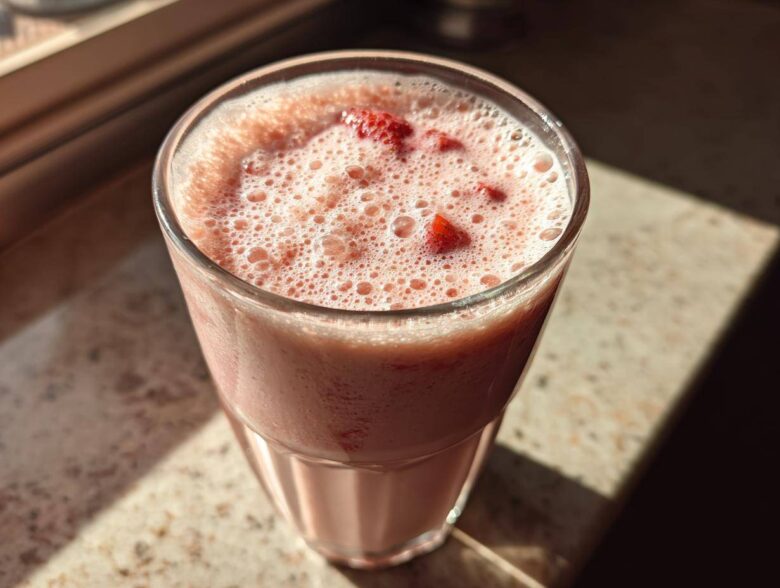 Close-up of a frothy Iced Vanilla Strawberry Protein Shake topped with small strawberry pieces in a clear glass.