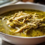Close-up of a white bowl filled with rich, green broth and shredded chicken from Instant Pot Chicken Pozole Verde.