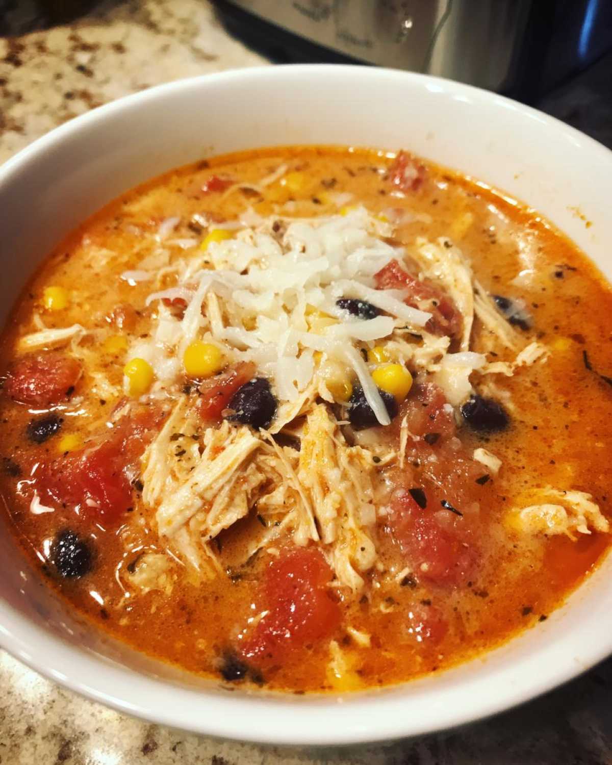 Close-up of a white bowl filled with Instant Pot Creamy Chicken Enchilada Soup, topped with shredded cheese, chicken, corn, and black beans.