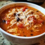 Close-up of a white bowl filled with Instant Pot Creamy Chicken Enchilada Soup, topped with shredded cheese, chicken, and black beans.