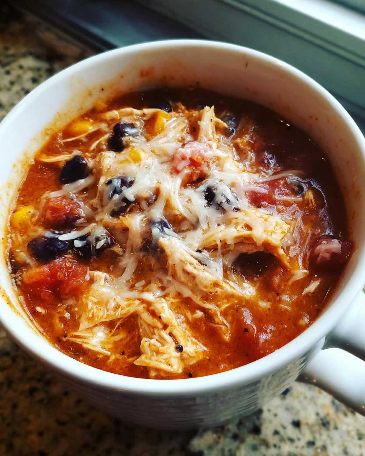A close-up of Instant Pot Creamy Chicken Enchilada Soup topped with melted shredded cheese, black beans, and corn.
