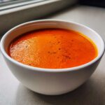 Close-up of a white bowl filled with vibrant orange Instant Pot Creamy Red Pepper Tomato Soup, garnished with pepper.