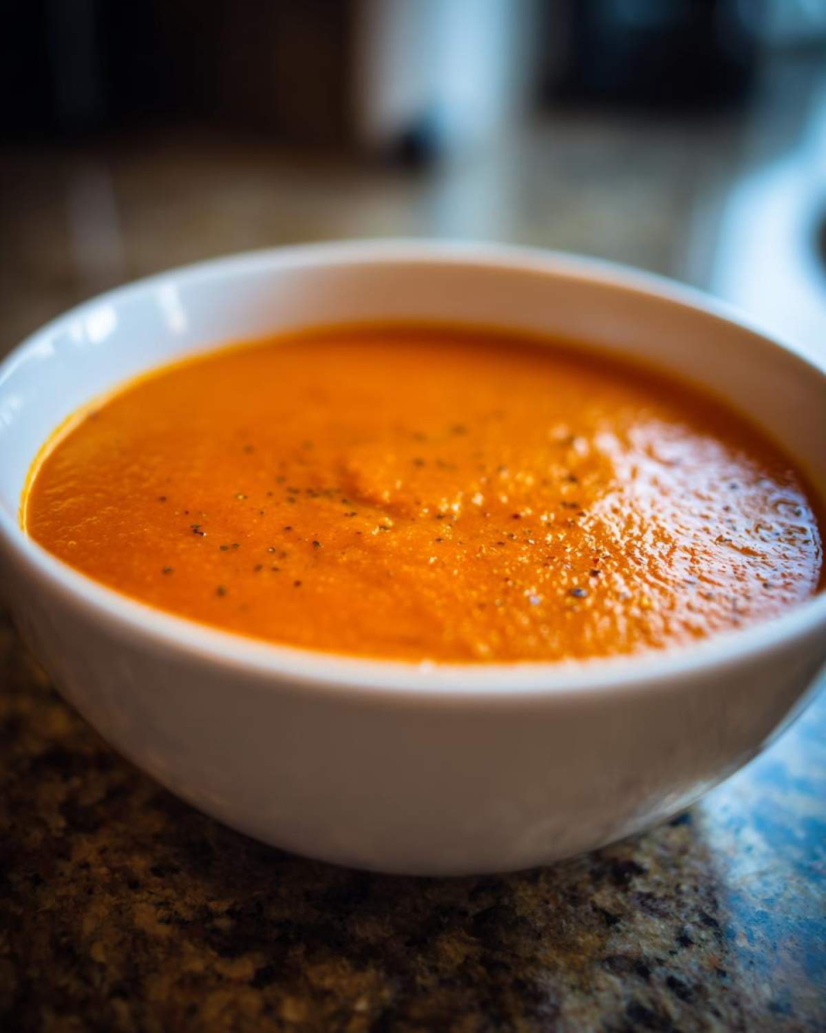 Close-up of a white bowl filled with vibrant orange Instant Pot Creamy Red Pepper Tomato Soup, topped with black pepper.