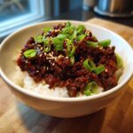 A close-up of a white bowl filled with white rice topped generously with saucy Instant Pot Korean Beef and garnished with green onions and sesame seeds.