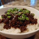 A white bowl filled with white rice topped with saucy Instant Pot Korean Beef, garnished with green onions and sesame seeds.