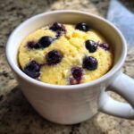 Close-up of a bright yellow Keto Blueberry Lemon Mug Cake topped with whole blueberries inside a white mug.