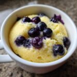A freshly baked Keto Blueberry Lemon Mug Cake, bright yellow and topped with whole blueberries, served in a white mug.