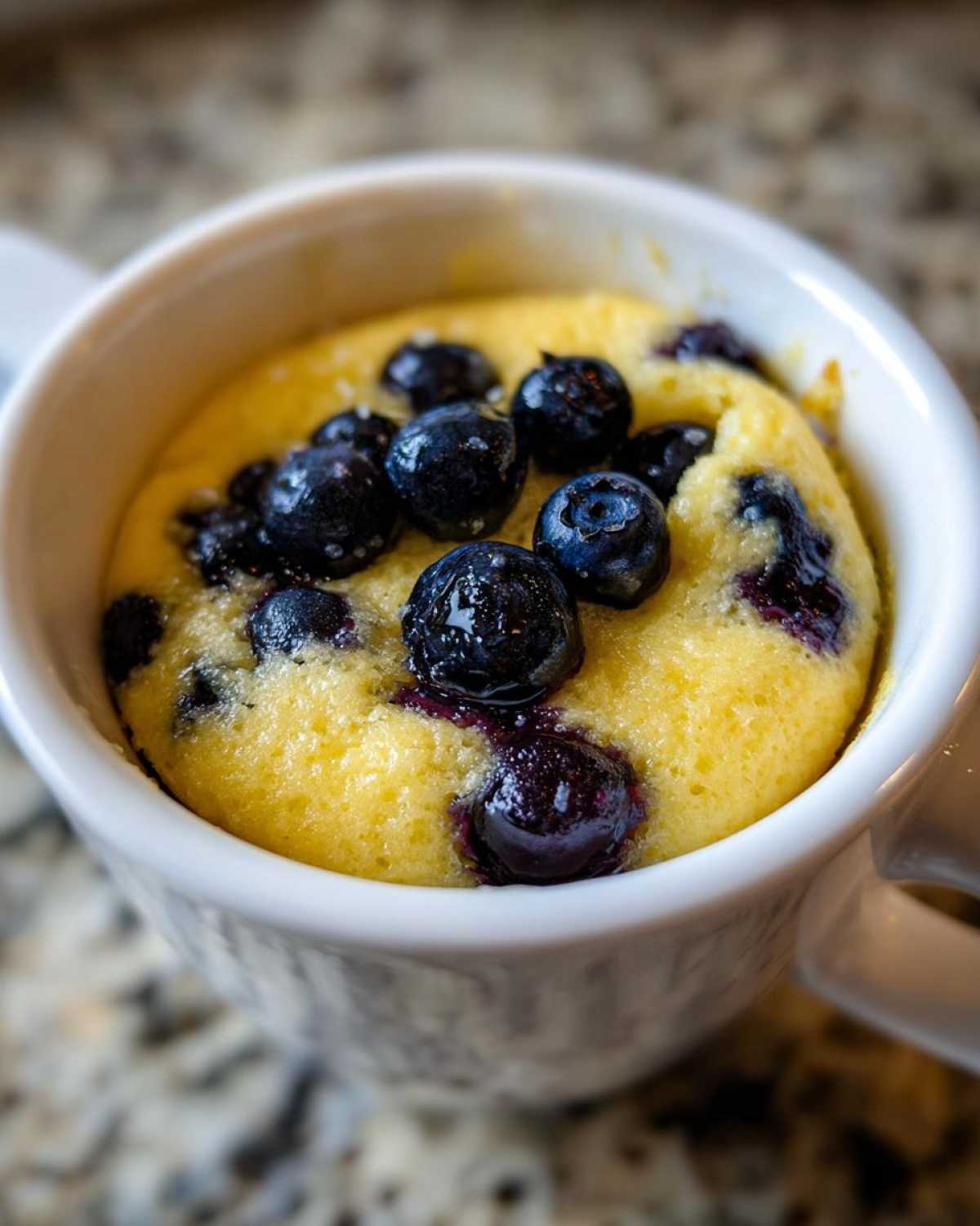 Close-up of a freshly baked Keto Blueberry Lemon Mug Cake topped with whole blueberries inside a white mug.