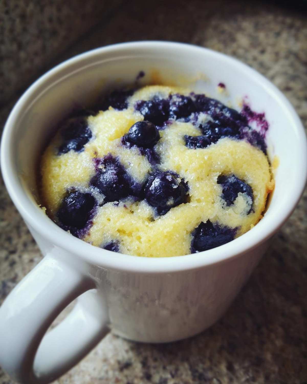 Close-up of a freshly baked Keto Blueberry Lemon Mug Cake, yellow cake studded with dark blueberries, served in a white mug.