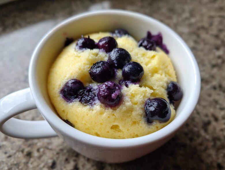 A freshly baked Keto Blueberry Lemon Mug Cake, bright yellow and topped with whole blueberries, served in a white mug.