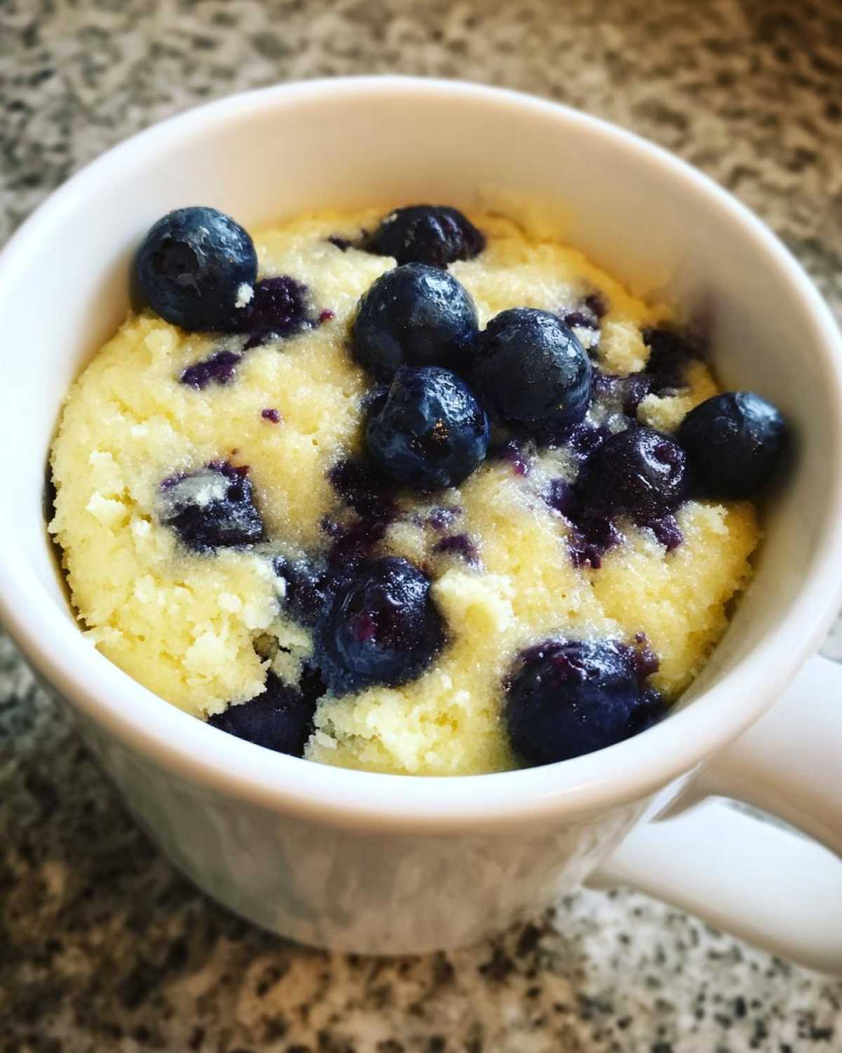 Close-up of a freshly baked Keto Blueberry Lemon Mug Cake topped with whole blueberries inside a white mug.