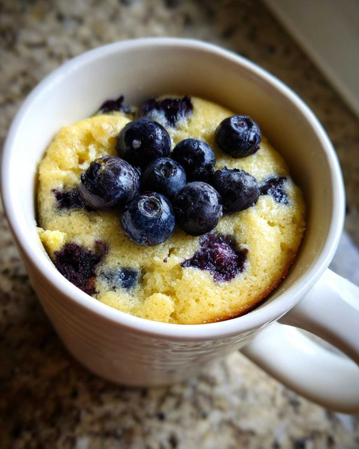 Close-up of a freshly baked Keto Blueberry Lemon Mug Cake topped with whole blueberries in a white ceramic mug.