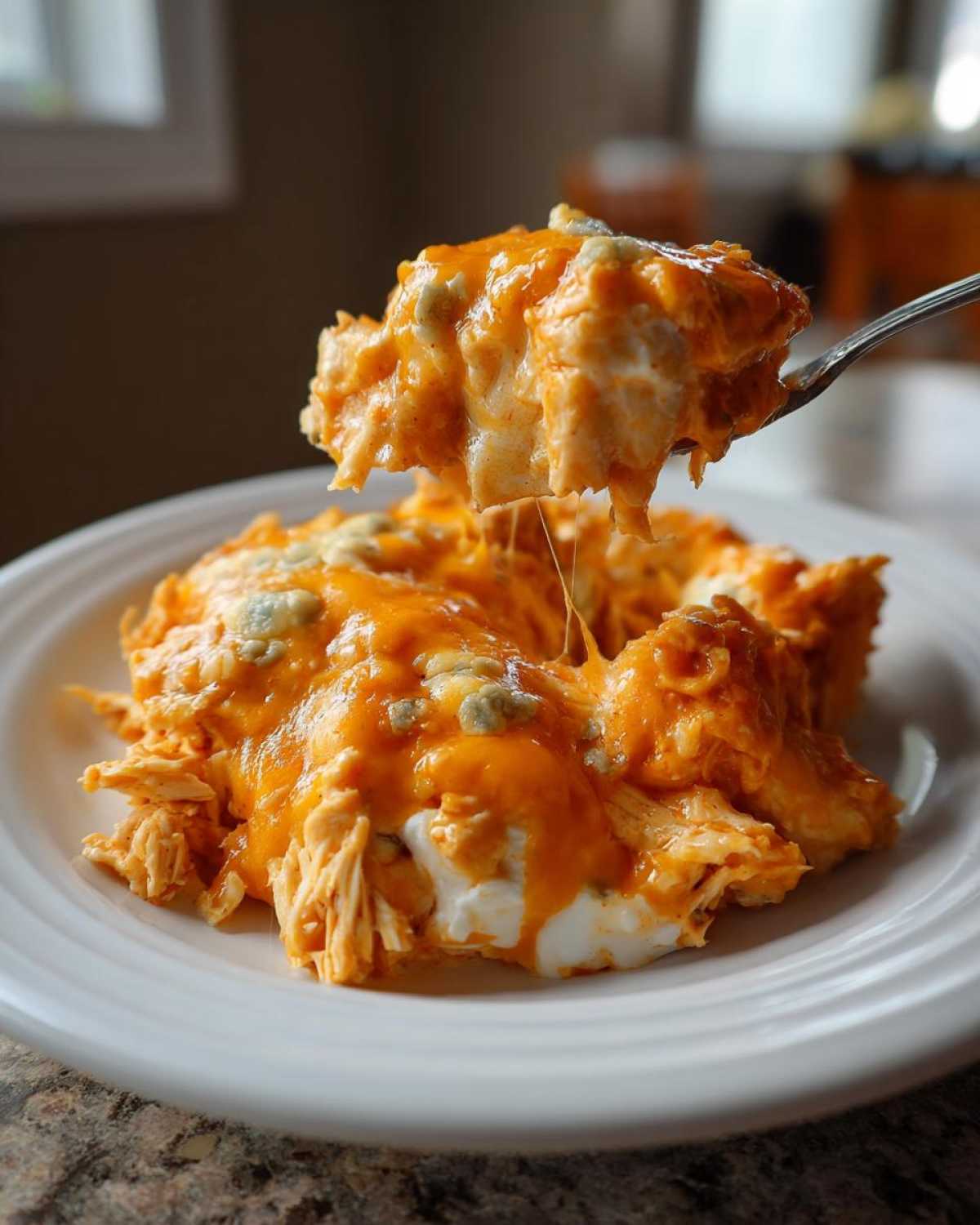 A spoonful of cheesy Keto Creamy Buffalo Chicken Casserole being lifted from a white plate, showing melted cheddar and blue cheese.