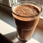 A tall glass filled with a thick, frothy Keto Creamy Chocolate Peanut Butter Shake sitting on a sunlit windowsill.