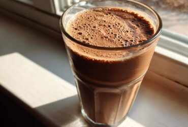 A tall glass filled with a thick, frothy Keto Creamy Chocolate Peanut Butter Shake sitting on a sunlit windowsill.