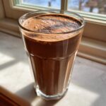 A tall glass filled with a thick, rich Keto Creamy Chocolate Peanut Butter Shake, sitting near a sunlit window.
