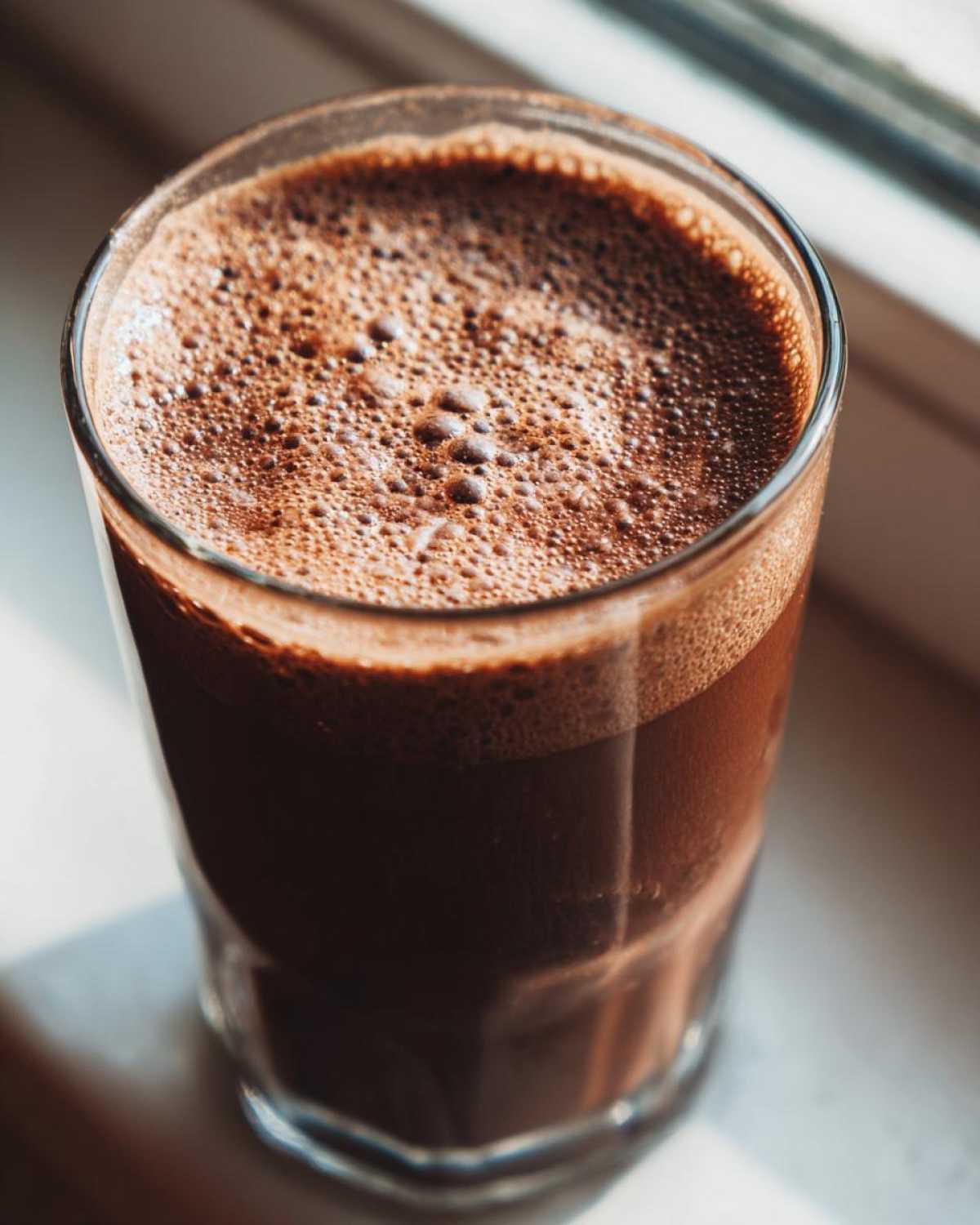 Close-up of a rich, dark Keto Creamy Chocolate Peanut Butter Shake with a thick layer of foam, sitting on a windowsill.