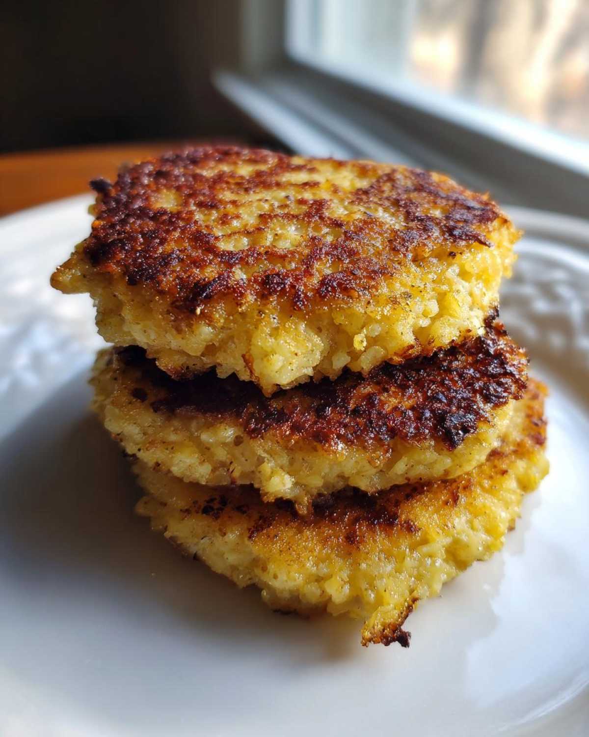 A stack of three golden-brown, crispy Keto Crispy Cauliflower Hash Browns on a white plate.