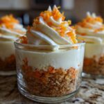 A close-up shot of a layered No-Bake Carrot Cake Cheesecake Cup featuring a graham cracker crust, creamy filling, and cream cheese frosting topped with orange zest.