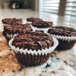 Several No-Bake Chocolate Coconut Protein Cups in white liners on a marble counter, drizzled with chocolate.