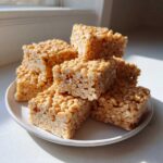 A stack of homemade No-Bake Peanut Butter Rice Cereal Bars piled on a white plate, glistening in sunlight.
