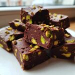 A stack of rich, dark chocolate No-Bake Pistachio Chocolate Fudge Squares topped with bright green pistachio pieces.