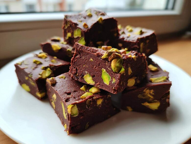 A stack of rich, dark chocolate No-Bake Pistachio Chocolate Fudge Squares topped with bright green pistachio pieces.