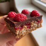 A hand holding a square piece of No-Bake Raspberry Chocolate Oat Slice showing layers of oats, raspberry jam, chocolate, and fresh raspberries.