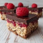 Close-up of a No-Bake Raspberry Chocolate Oat Slice showing oat base, raspberry jam, and chocolate topping with a fresh raspberry.