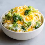 A close-up of a white bowl filled with One-Pot Creamy Broccoli Cheddar Rice, topped with melted cheddar cheese.